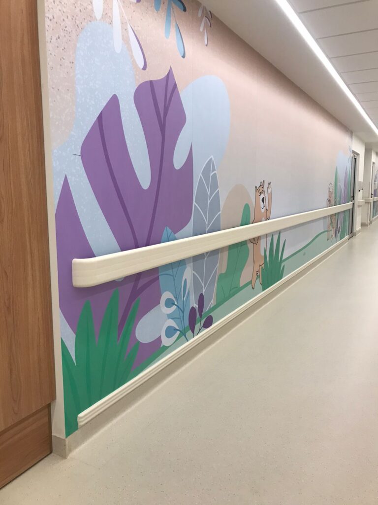 A imagem mostra um corredor com um design voltado para um ambiente infantil. A parede apresenta um mural colorido com ilustrações de folhas, plantas e pequenos animais. Há protetores de parede branco instalado horizontalmente, servindo tanto para proteção quanto como apoio para circulação.